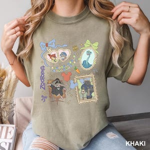 Puede incluir: Una camiseta color caqui con un estampado gr&aacute;fico de Zootopia que incluye personajes como Nick Wilde, Judy Hopps, Mr. Big, Flash y el Jefe Bogo en marcos ornamentados, junto a una placa de polic&iacute;a, una zanahoria y globos con forma de Mickey.