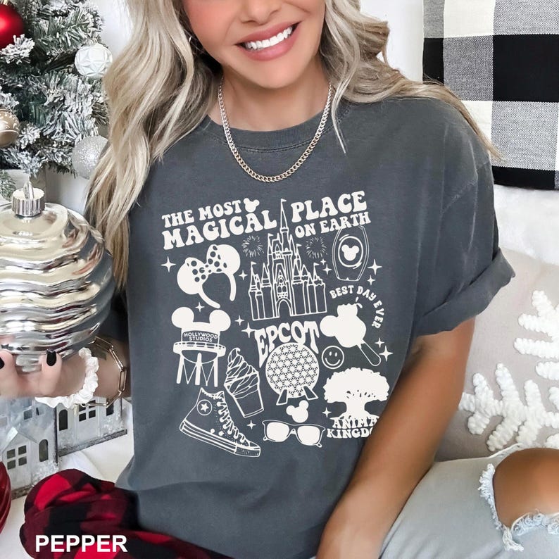 Puede incluir: Camiseta gris oscuro con un dise&ntilde;o gr&aacute;fico blanco que presenta el texto "THE MOST MAGICAL PLACE ON EARTH" y varios iconos de los parques de Disney, incluyendo un castillo, orejas de Mickey Mouse y un helado. La camiseta tiene mangas cortas y un ajuste relajado.