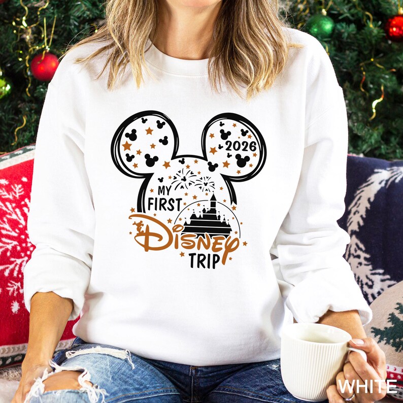 May include: White sweatshirt with a black and brown Mickey Mouse design. The design includes the text "MY FIRST DISNEY TRIP" and the year "2026". The sweatshirt is a crew neck style.