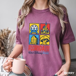Może przedstawiać: Koszulka w kolorze jagodowym z kolorową grafiką czterech kwadrat&oacute;w z kultowymi zabytkami Florydy. Tekst "FLORIDA!!! Walt Disney World" jest wydrukowany poniżej. Koszulka ma wygodny kr&oacute;j.