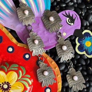 May include: Close-up of silver-toned earrings with a bird design and a pink center stone. The earrings are hanging in front of a red and purple painted background with a floral design. The background is surrounded by black beans.