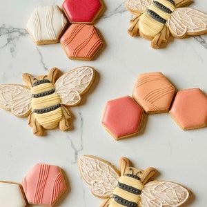 May include: Assortment of decorated sugar cookies featuring bees and honeycomb designs. The cookies are decorated with pink, white, and orange icing.