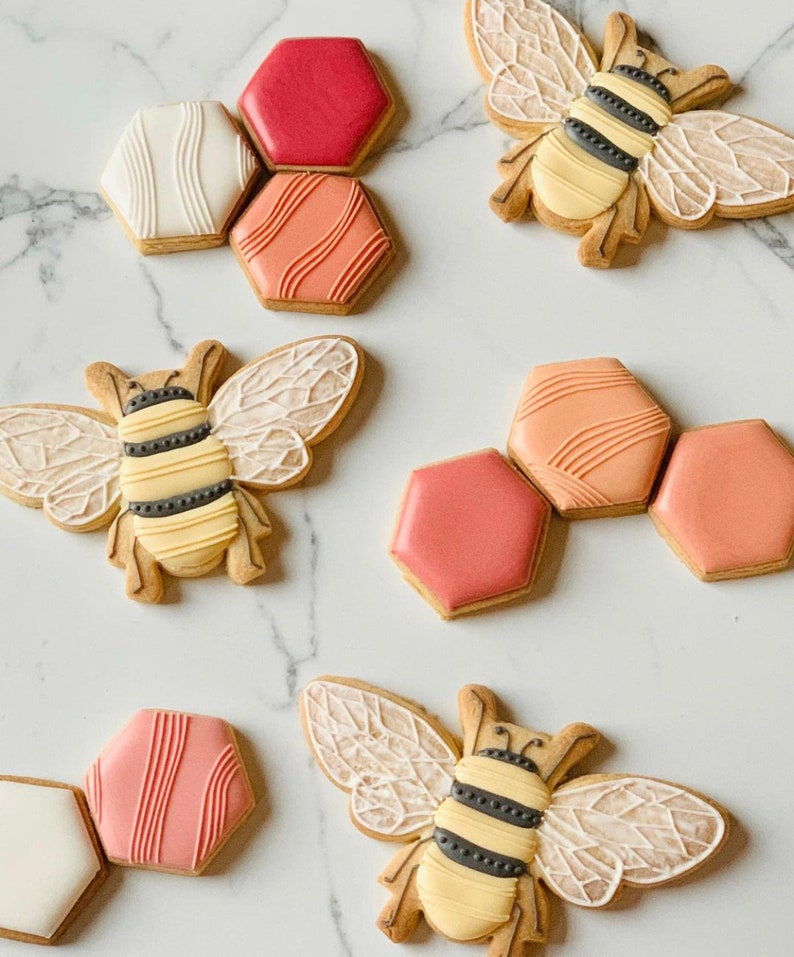 May include: Assortment of decorated sugar cookies featuring bees and honeycomb designs. The cookies are decorated with pink, white, and orange icing.