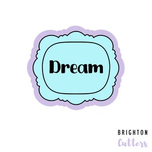 May include: A light blue cookie cutter with a scalloped edge and the word "Dream" in black text. The cookie cutter is on a white background with the text "Brighton Cutters" in purple text below.