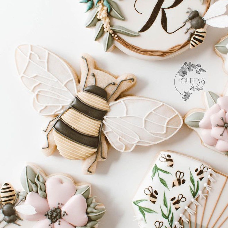 May include: A collection of decorated sugar cookies featuring a bee, flowers, and a beehive design. The cookies are decorated with royal icing and have a delicate, whimsical feel.