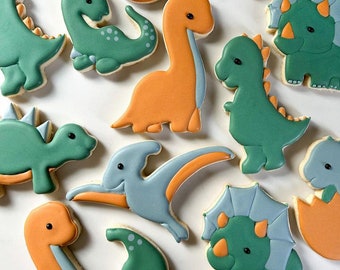 May include: A collection of decorated sugar cookies shaped like dinosaurs. The cookies are decorated with green, blue, orange, and brown icing. The cookies are arranged on a white surface.