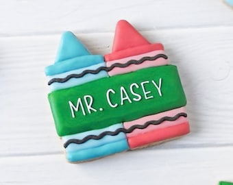 Op de afbeelding: Een koekje in de vorm van twee naast elkaar liggende kleurpotloden, één lichtblauw en één roze, met een groene middenband waarop de witte tekst MR. CASEY staat. Het koekje is versierd met zwarte golvende lijnen en glad glazuur.