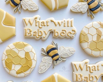 May include: A set of nine decorated sugar cookies with a bee and honeycomb theme. The cookies are decorated with white icing and yellow and gold accents. The text "What will baby bee?" is written on two of the cookies.