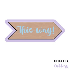 May include: A wooden arrow sign with a light purple border. The sign says "This way!" in light blue cursive text.