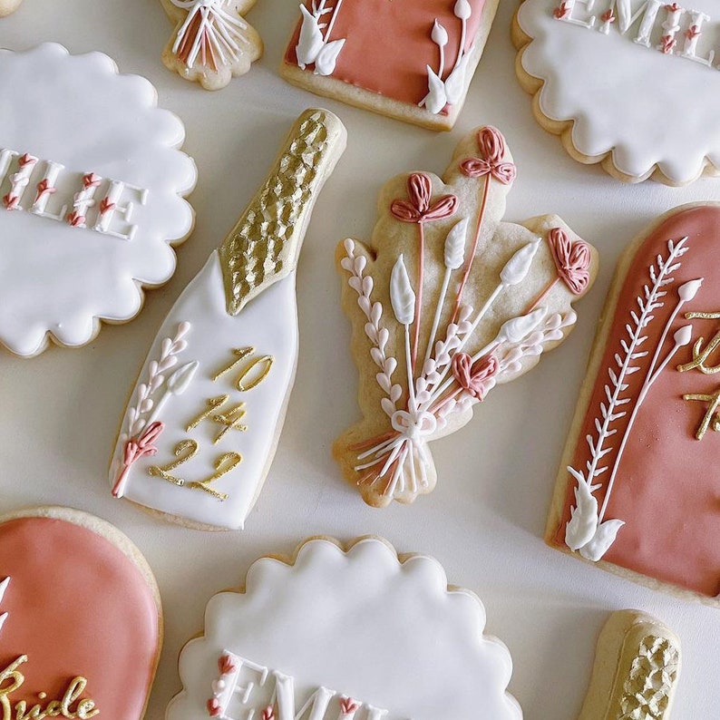 May include: Assortment of decorated sugar cookies with various designs, including a champagne bottle with the date 10.14.22, a bouquet of flowers, and the word "Emily" written on several cookies.