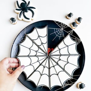 May include: A black plate with a white spiderweb design, surrounded by spider-shaped cookies. The cookies are decorated with black and white icing. A large spider cookie is also present.