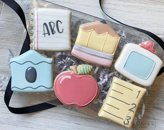 Op de afbeelding: Een set van zes school-thema suikerkoekjes in een doorzichtige doos, inclusief een notitieboek met ABC, een potlood, een lijmfles, een lichtblauw object, een rode appel met een groen blaadje en een gele liniaal met de cijfers één tot drie.