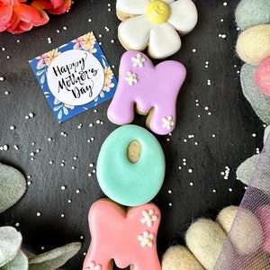 May include: Decorated sugar cookies spelling out "MOM" with a flower cookie on top. The cookies are decorated with pastel pink, purple, and blue icing and white flowers. A card with the text "Happy Mother's Day" is in the background.