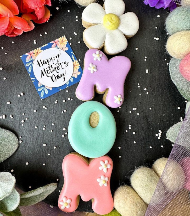 May include: Decorated sugar cookies spelling out "MOM" with a flower cookie on top. The cookies are decorated with pastel pink, purple, and blue icing and white flowers. A card with the text "Happy Mother's Day" is in the background.