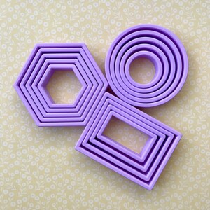 May include: Set of four purple plastic cookie cutters. The cutters are in the shapes of a rectangle, a circle, and a hexagon. The hexagon cutter is made up of three concentric rings.