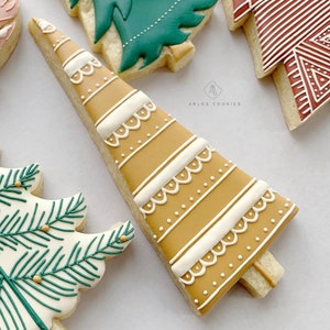 May include: A gingerbread cookie decorated with white and gold icing in the shape of a Christmas tree. The cookie has a white lace pattern and gold dots.