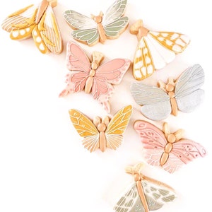 May include: A collection of butterfly-shaped cookies with intricate details. The cookies feature a variety of colors, including pink, yellow, green, and white, with delicate icing designs. The cookies are arranged on a white surface.