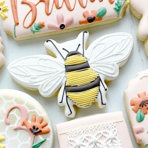 May include: A white and yellow bee cookie with black stripes and white wings. The cookie is decorated with icing and is on a white surface.