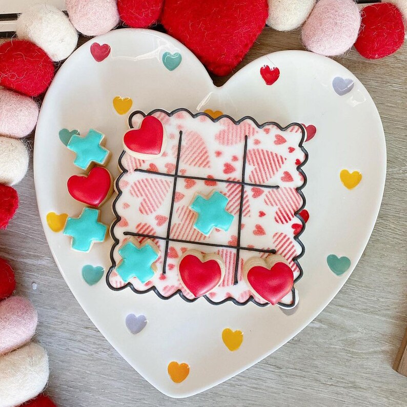 K&ouml;nnte beinhalten: Ein herzf&ouml;rmiger Teller mit einem Tic-Tac-Toe-Keks, der mit roten und t&uuml;rkisfarbenen Herzen verziert ist. Der Teller ist mit kleinen Herzen in verschiedenen Farben verziert.