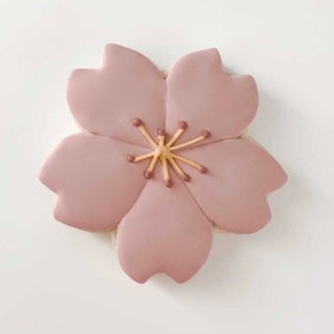 May include: A pink sugar cookie decorated with a five-petal flower design. The flower has a brown centre with a yellow stamen.
