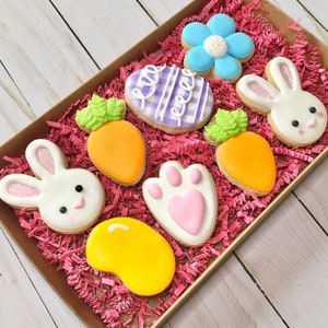 May include: A box filled with pink crinkle paper and decorated Easter cookies. The cookies are shaped like a bunny, a paw print, a carrot, a flower, an egg, and a jelly bean.