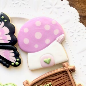 May include: A set of three decorated sugar cookies. One cookie is shaped like a butterfly with black and pink wings. Another cookie is shaped like a mushroom with a pink and white top and a white base. The third cookie is shaped like a wooden sign with brown and white paint.