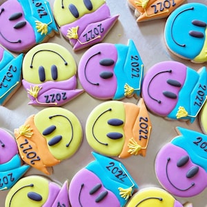 May include: Assortment of colourful decorated sugar cookies with smiley faces and the year 2022. The cookies are decorated with blue, yellow, orange, and purple icing.