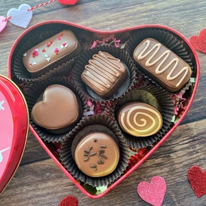 May include: A red heart-shaped box filled with assorted chocolate candies. The box is open and the candies are visible. The candies are in various shapes and sizes, including hearts, squares, and swirls. The candies are all brown and have a chocolatey appearance.