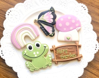 Op de afbeelding: Vijf gedecoreerde suikerkoekjes op een wit bord met een kantrand. De koekjes zijn gevormd als een regenboog, een kikker, een vlinder, een paddenstoel en een houten bord met de tekst "Fairy Garden".