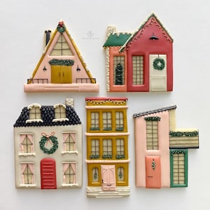 Puede incluir: Una variedad de galletas de jengibre decoradas con forma de casas, en varias formas y colores. Las galletas presentan glaseado rosa, rojo, amarillo y blanco, con detalles como coronas, ventanas y puertas. Algunas tienen detalles verdes y blancos.