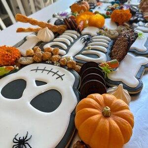 May include: A Halloween-themed cookie platter featuring a skeleton cookie with a skull, surrounded by various treats like mini pumpkins, candy corn, pretzels, and chocolate cookies.