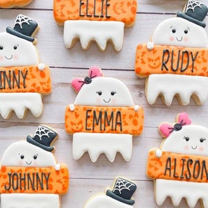 May include: A selection of Halloween ghost-shaped biscuits, each with a name on an orange banner. The biscuits feature white icing, black hats, and pink bows. Names include "Ellie", "Rudy", "Emma", "Alison", and "Johnny".