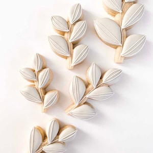 Peut inclure: Cinq biscuits sablés blancs décorés de glaçage blanc en forme de feuilles stylisées. Les biscuits sont disposés sur une surface blanche.