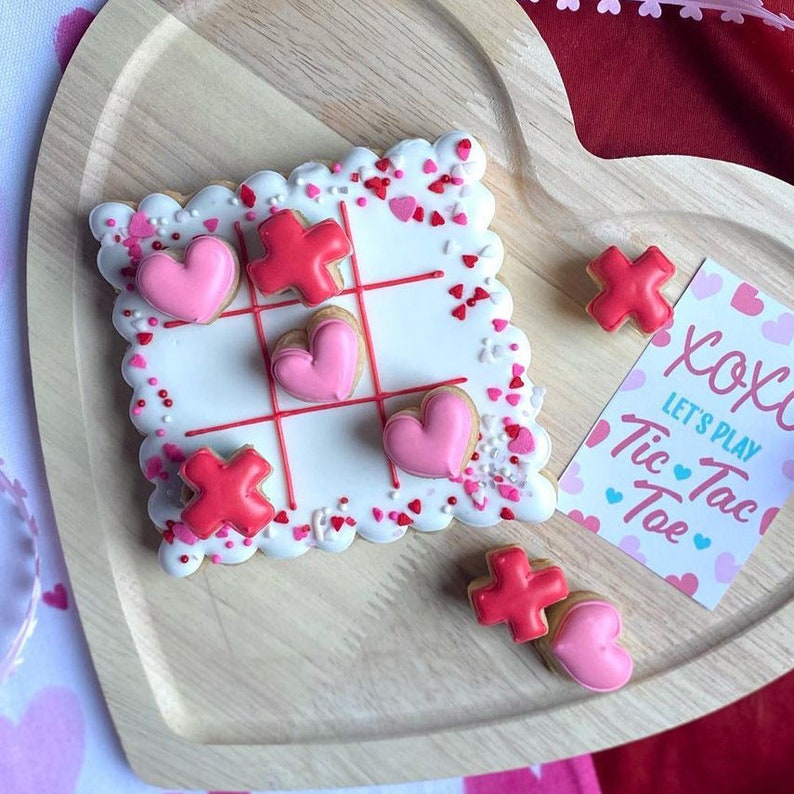 K&ouml;nnte beinhalten: Ein wei&szlig;er Keks, der mit rosa Herzen und roten Kreuzen verziert ist und einem Tic-Tac-Toe-Spielfeld &auml;hnelt. Der Keks ist von rosa und roten Streuseln umgeben. Eine separate Karte lautet "Lass uns Tic Tac Toe spielen" mit rosa Herzen und blauem Text.