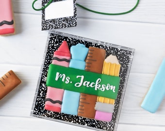Op de afbeelding: Een vierkant koekje versierd met glazuur in de vorm van schoolspullen zoals een kleurpotlood, lijm, liniaal en potlood, met een groene banner met de witte tekst Ms. Jackson. Een bijpassend cadeaulabel met groen touwtje is inbegrepen.
