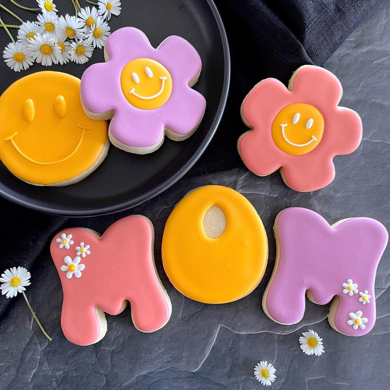 May include: A set of decorated sugar cookies spelling out "MOM" with yellow, pink, and purple frosting. The cookies are decorated with smiley faces and flowers.