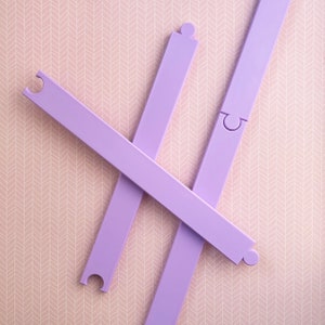 Puede incluir: Tres piezas de rompecabezas de plástico morado con bordes que se entrelazan. Las piezas están dispuestas en forma de cruz sobre un fondo rosa.