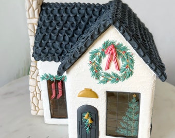 May include: A white gingerbread house with a black shingled roof, a green wreath with a red ribbon, and a gold bell on the door. The windows are decorated with green Christmas trees and red stockings.
