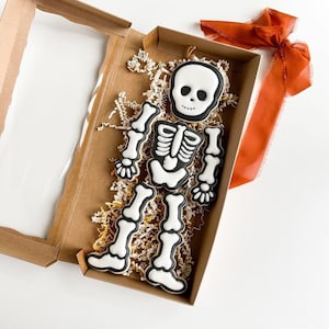 May include: A skeleton cookie set in a brown box with white tissue paper. The skeleton is made of black icing with white details. The cookies are in the shape of a head, torso, and two arms and legs.