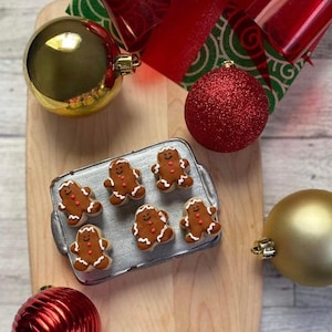 May include: A silver baking pan with six gingerbread men cookies decorated with white icing and red candy dots. The pan is on a wooden surface with red and gold ornaments.