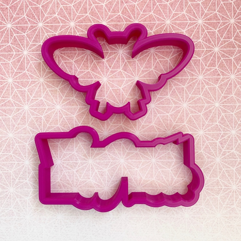 May include: Two pink plastic cookie cutters, one shaped like a butterfly and the other shaped like a banner.