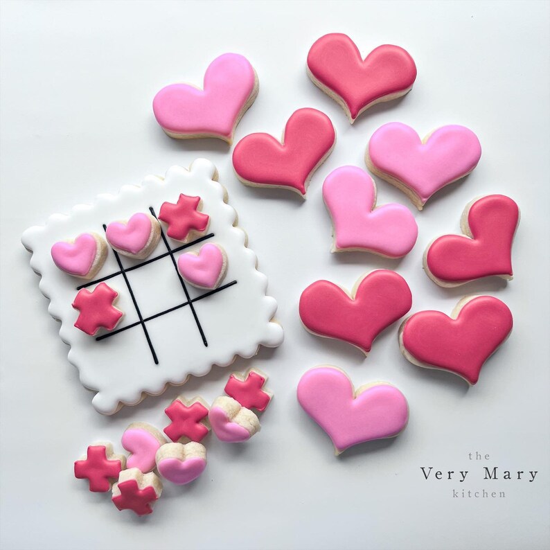 K&ouml;nnte beinhalten: Ein Set aus rosafarbenen und roten herzf&ouml;rmigen Keksen, einige mit einem Tic-Tac-Toe-Brett-Design und andere mit einer einfachen Herzform. Die Kekse sind mit wei&szlig;em Zuckerguss und schwarzen Linien verziert. Der Text "the Very Mary kitchen" ist am unteren Rand des Bildes sichtbar.