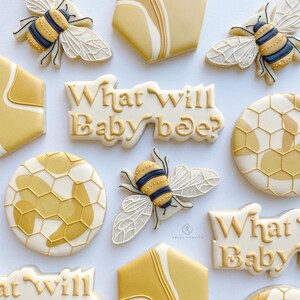 May include: A set of nine decorated sugar cookies with a bee and honeycomb theme. The cookies are decorated with white icing and yellow and gold accents. The text "What will baby bee?" is written on two of the cookies.