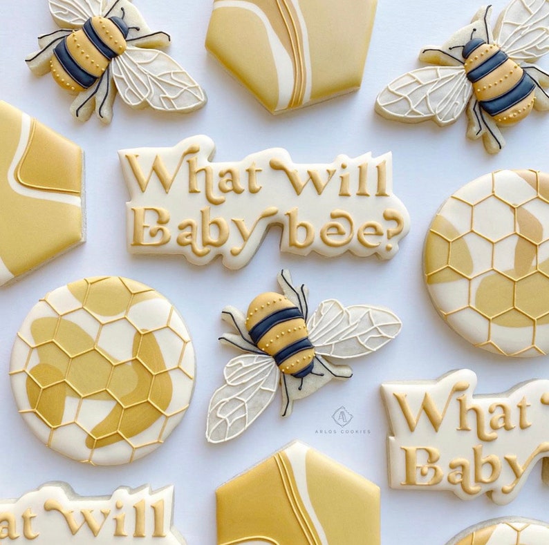 May include: A set of nine decorated sugar cookies with a bee and honeycomb theme. The cookies are decorated with white icing and yellow and gold accents. The text "What will baby bee?" is written on two of the cookies.