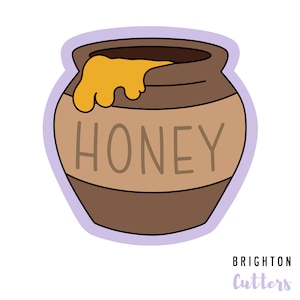 Peut inclure: Un pot de miel en céramique brun avec un filet de miel jaune et le mot "HONEY" écrit sur le devant. Le pot est bordé de noir et a un fond violet.