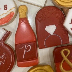 May include: Assortment of red, white, and gold decorated cookies. The cookies are shaped like a champagne bottle, a ring with a diamond, an arch with a design, and a circle with an ampersand. The cookies are decorated with royal icing.