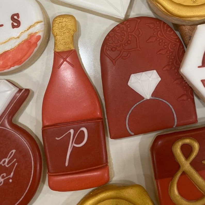 May include: Assortment of red, white, and gold decorated cookies. The cookies are shaped like a champagne bottle, a ring with a diamond, an arch with a design, and a circle with an ampersand. The cookies are decorated with royal icing.