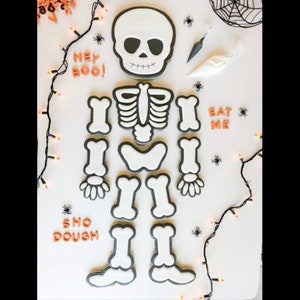 May include: A set of Halloween skeleton cookies decorated with black icing. The cookies are arranged to form a skeleton with the words "Hey Boo!" and "Eat Me" on the top and bottom of the skeleton. The cookies are on a white background with a string of lights and black spiders.