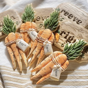May include: Three decorated sugar cookies shaped like carrots with green tops. Each carrot has a white tag with a name written on it: Reeve, Riggin, and Reeve.
