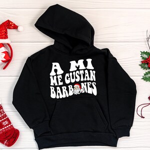 Op de afbeelding: Zwart hoodie met een witte afbeelding die "A MI ME GUSTAN BARBONES" zegt met een cartoon kerstman in het midden van de tekst.